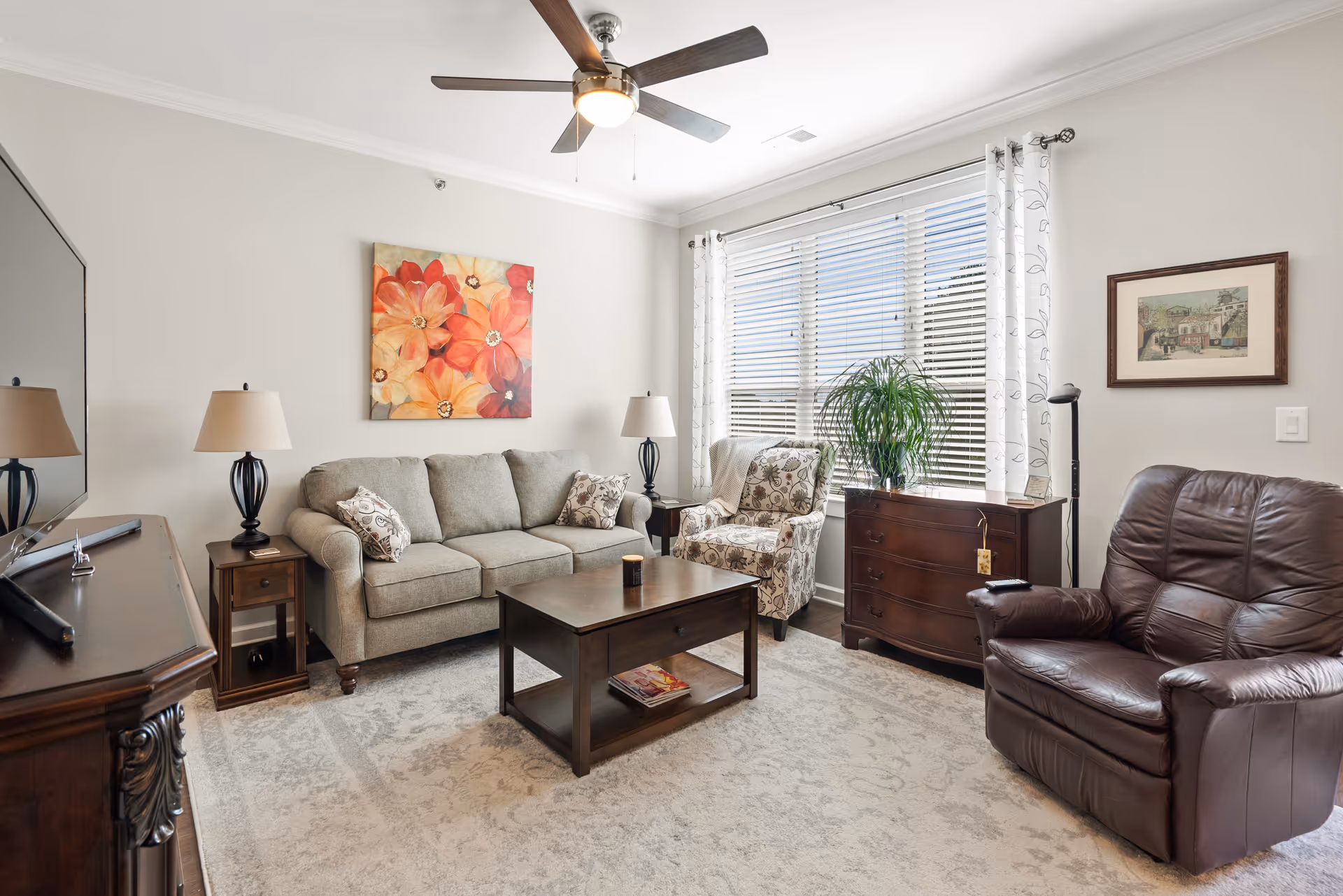 A cozy living room with a gray sofa, a patterned armchair, and a brown leather recliner. There is a wooden coffee table in the center with a candle on it, a wooden side table with a lamp, and a wooden dresser with a green plant on top. The room has a large window with white blinds and sheer curtains, a ceiling fan with a light, and a colorful floral painting on the wall.