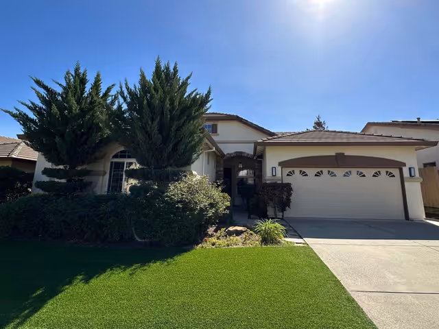 Front exterior of a single-family style building with a two-car garage, driveway, manicured lawn, and trees under a clear blue sky.