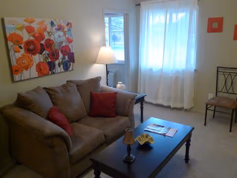 A cozy living room with a brown sofa adorned with red cushions, a dark wooden coffee table with a small lamp and decorative bowl, a side table with a lamp, a window with sheer white curtains, a chair against the wall, and a colorful floral painting hanging above the sofa.