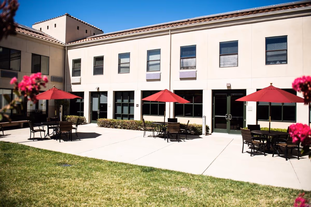 Outdoor patio area at The Reutlinger Community with several tables and chairs under red umbrellas, surrounded by a building with multiple windows and doors. There is a green lawn in the foreground and some pink flowers partially visible on the sides.