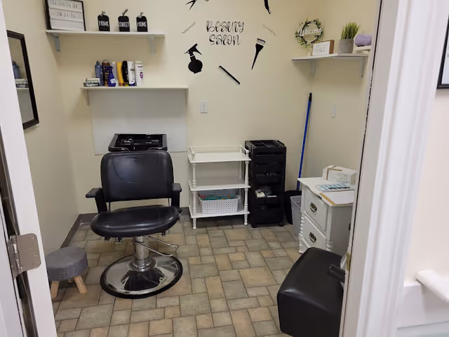 A small beauty salon room with a black salon chair in the center, a small gray stool to the left, shelves on the walls holding hair care products and decorative items, a white rolling cart with supplies, and wall decals depicting beauty tools and the words 'Beauty Salon'.