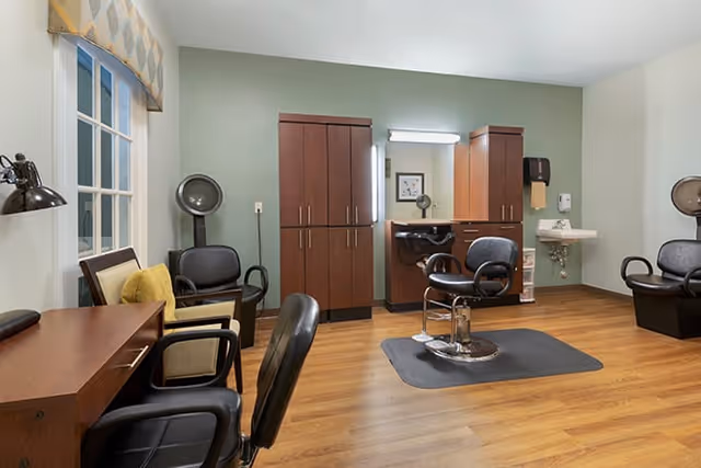 Interior of a hair salon room with wooden flooring, a styling chair on a mat in front of a mirror with cabinets, additional black chairs, a hair dryer, a small sink, and a window with a patterned valance.