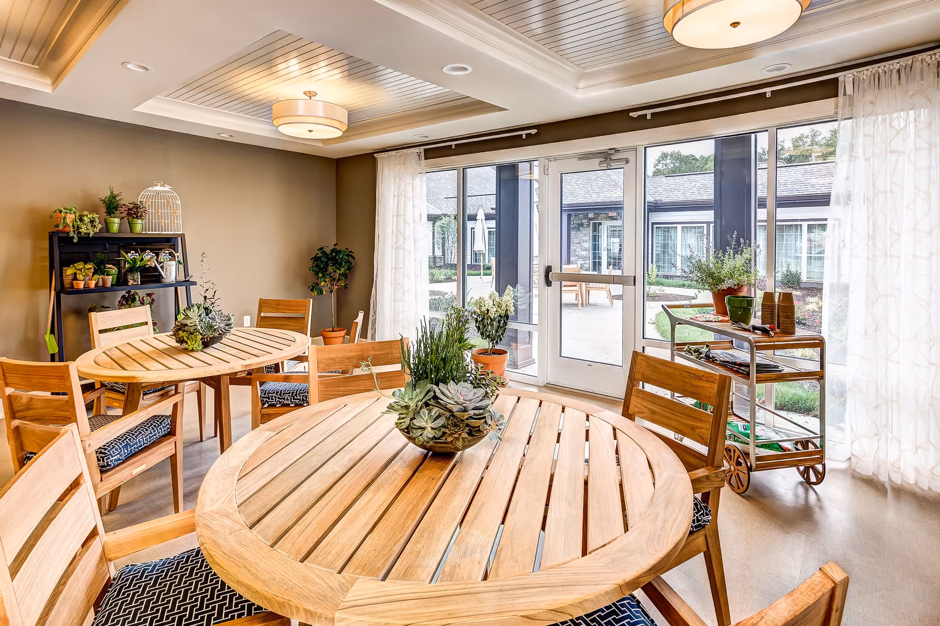 A bright and inviting common area with round wooden tables and chairs, decorated with potted plants and succulents. Large windows and a glass door provide a view of an outdoor patio area with additional seating. The room features a modern ceiling light fixture and sheer white curtains.