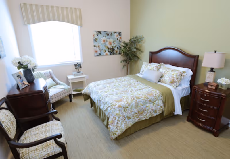 A cozy bedroom with a bed featuring floral bedding and multiple pillows. The room includes two wooden nightstands with lamps and decorative items, two upholstered chairs, a small white side table, a window with a striped valance, a floral wall painting, and a potted plant in the corner.