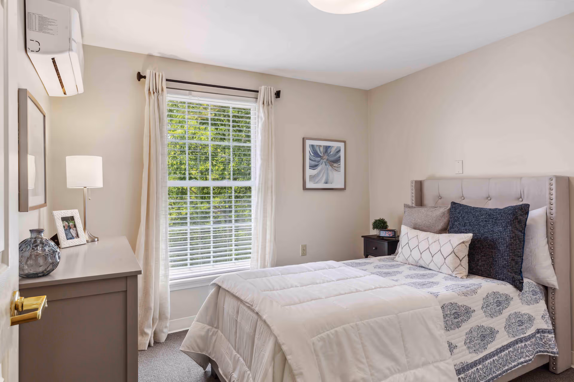 A cozy bedroom with a neatly made bed featuring a white comforter and patterned blue and white bedding. The bed has a tufted headboard with decorative pillows. To the right of the bed is a small dark nightstand with a small plant and a clock. A large window with white blinds and light beige curtains lets in natural light, and a framed abstract artwork hangs on the wall. On the left side, there is a dresser with a lamp, a framed photo, and a decorative vase. The walls are painted light beige, and the room has a calm, inviting atmosphere.