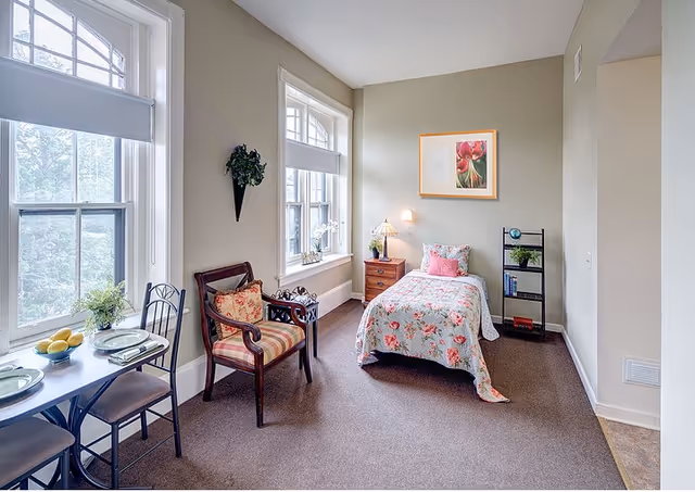 A bright and cozy bedroom with a single bed covered in floral bedding, a wooden nightstand with a lamp and plant, a small black bookshelf with books and a globe, a wooden armchair with a floral cushion, and a small dining table set for two near large windows with white blinds.