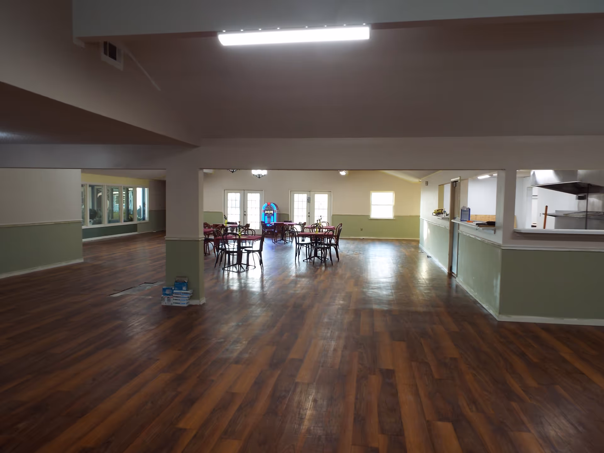 Interior view of a spacious dining area with wooden flooring, several round tables with chairs, a jukebox near the back wall, and a serving window to a kitchen on the right side.