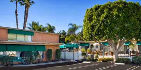 Outdoor view of a senior living facility with a single-story building featuring green awnings, palm trees, and a large leafy tree in the courtyard area under a clear blue sky.