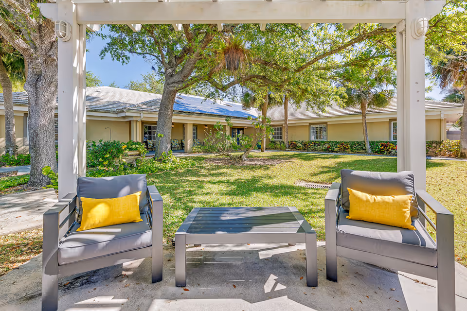 Outdoor seating area with two gray armchairs each with a yellow pillow and a matching gray coffee table under a white pergola, surrounded by green grass, trees, and a single-story building in the background.