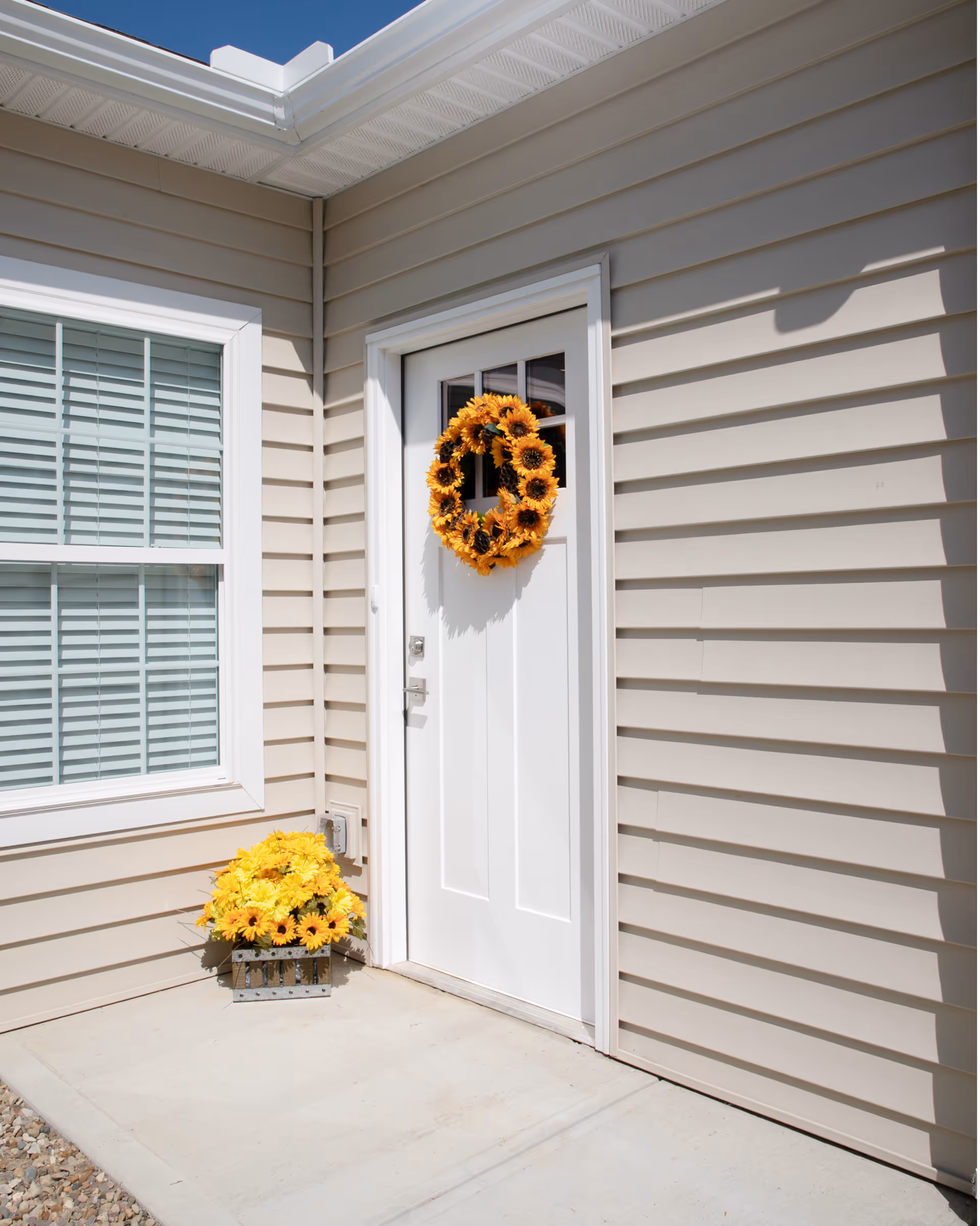 A white exterior door on beige siding decorated with a sunflower wreath and a matching planter of yellow flowers beside a window with blinds.