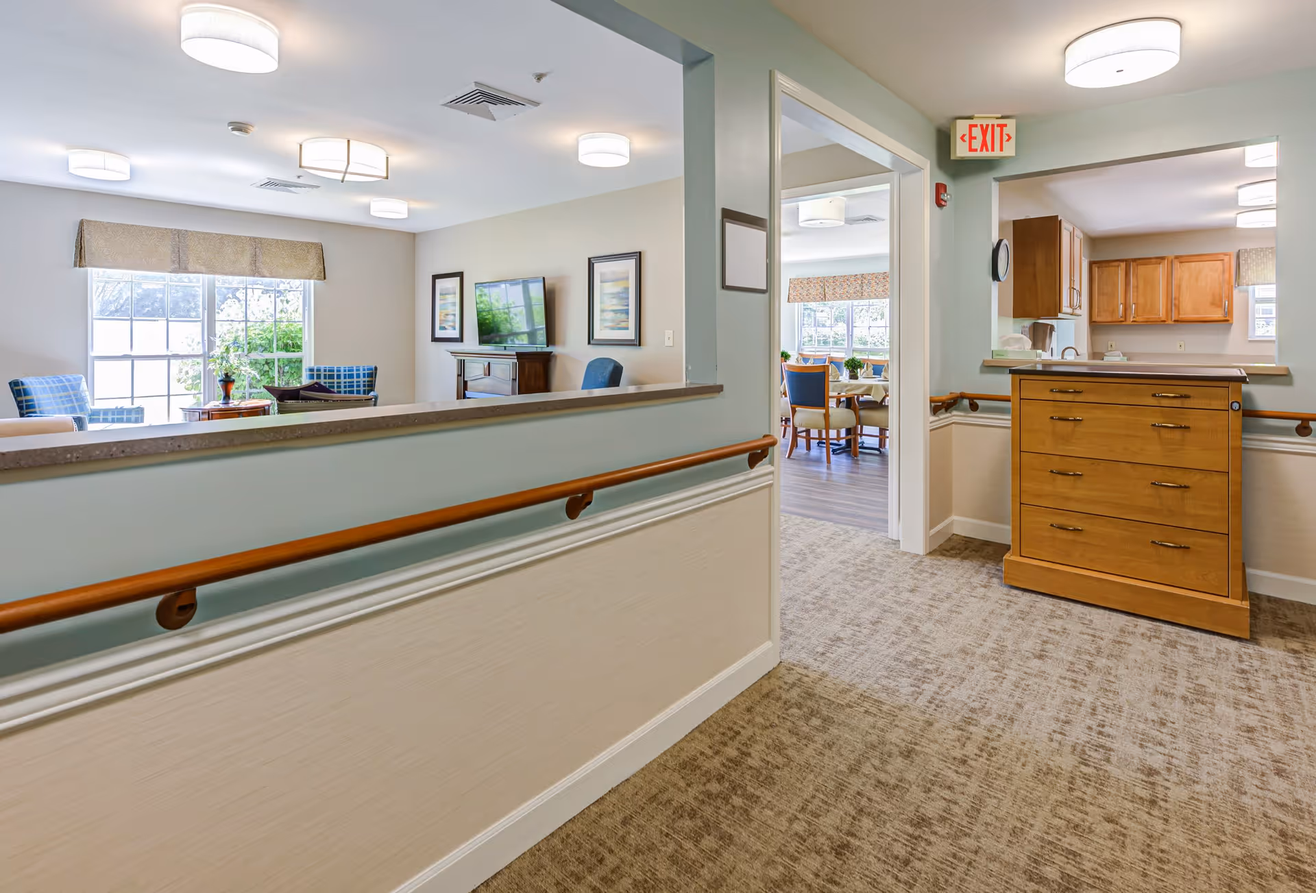 Interior view of a senior living facility hallway with beige walls and carpeted floor. The hallway has wooden handrails on both sides and leads to a living room area with blue chairs, a TV, and framed artwork on the walls. To the right, there is a kitchen area with wooden cabinets and a wooden drawer unit. An exit sign is visible above a doorway.