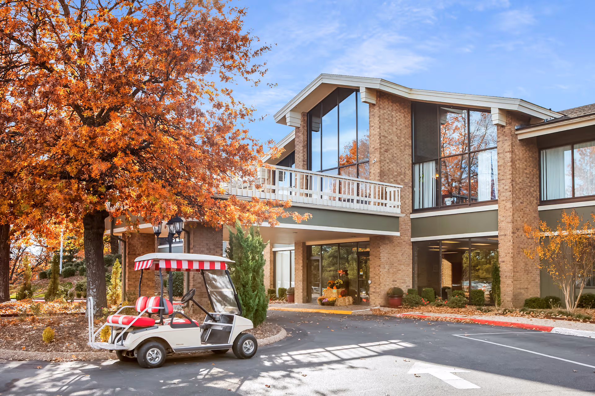 Front exterior of a brick senior living facility with a parked red-and-white-striped golf cart and autumn trees.