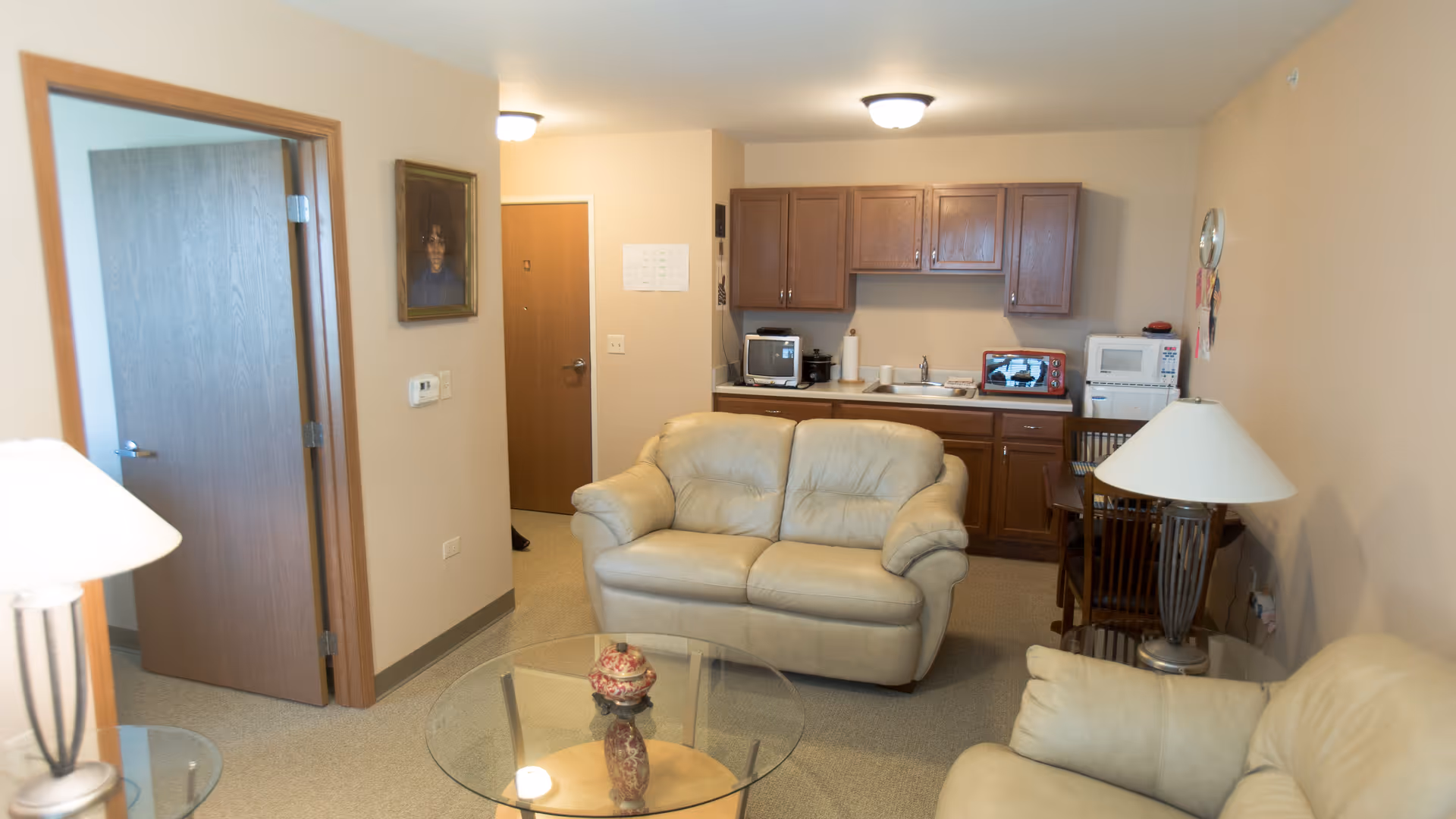 A small living area with two beige leather sofas facing a round glass coffee table with a decorative vase on it. In the background, there is a kitchenette with wooden cabinets, a microwave, a toaster oven, and a small dining table with chairs. A lamp with a white shade is visible on the right side, and an open door leads to another room on the left.