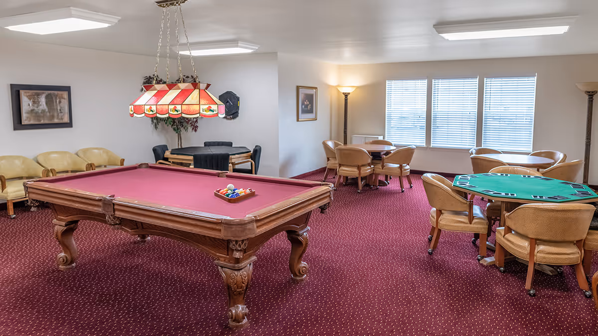 A recreational room with a red felt pool table in the center, surrounded by beige chairs and multiple round tables with chairs. The room has red carpet flooring, white walls, large windows with blinds, and two floor lamps providing light. A decorative hanging lamp is above the pool table.