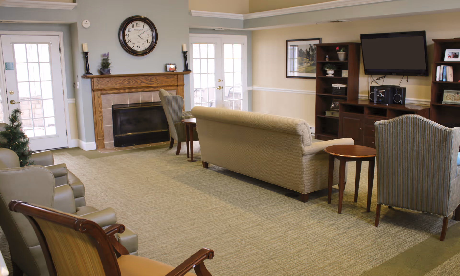 A cozy living room area in a senior living facility with a beige sofa, two armchairs, a wooden rocking chair, a fireplace with a clock above it, a TV mounted on the wall, and wooden shelves with decorative items. There are glass doors letting in natural light.