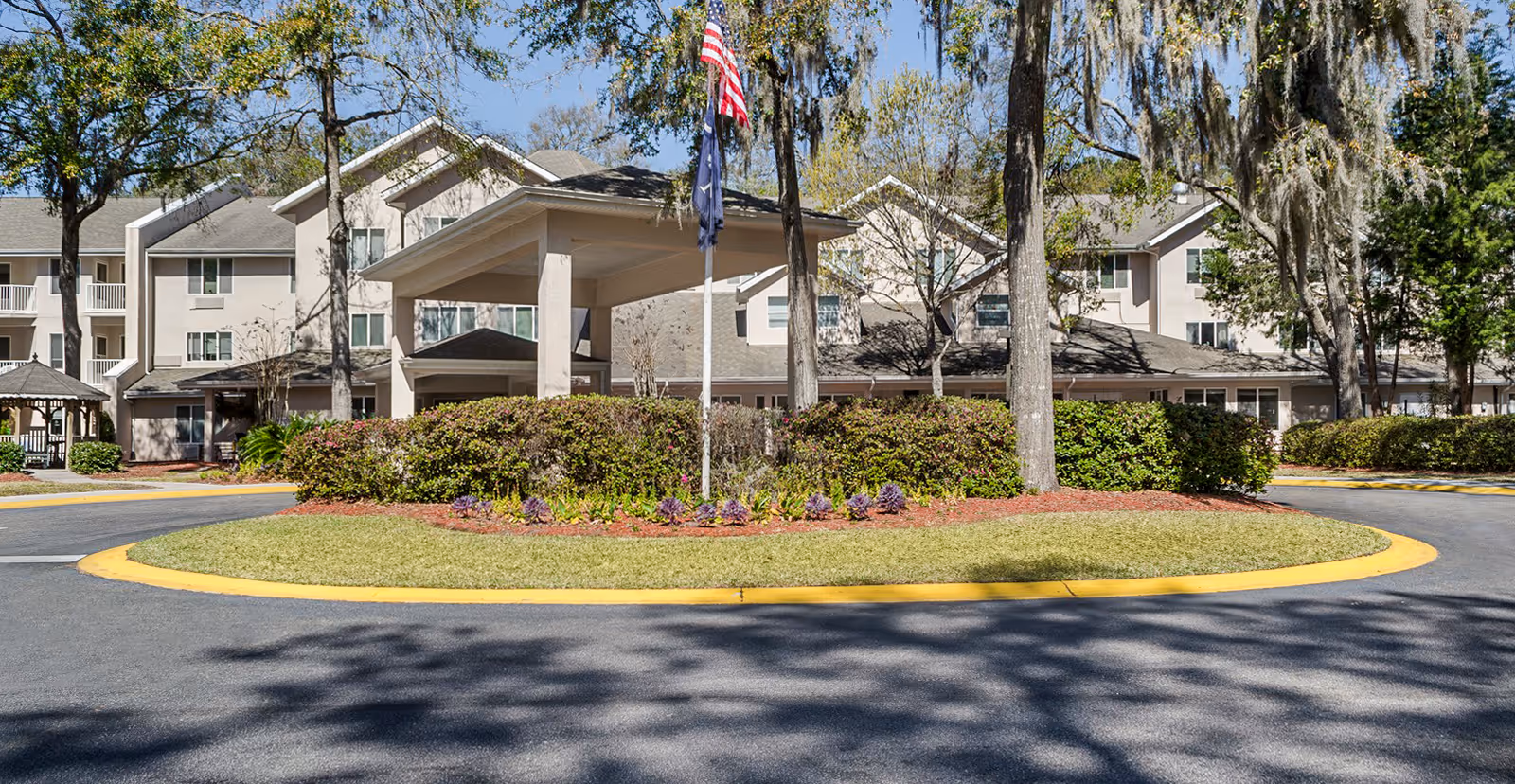 Exterior view of Holiday Indigo Pines facility showing a circular driveway with a landscaped island in the center, featuring bushes, flowers, and two flagpoles with flags. The building is a multi-story structure with balconies and surrounded by trees under a clear blue sky.