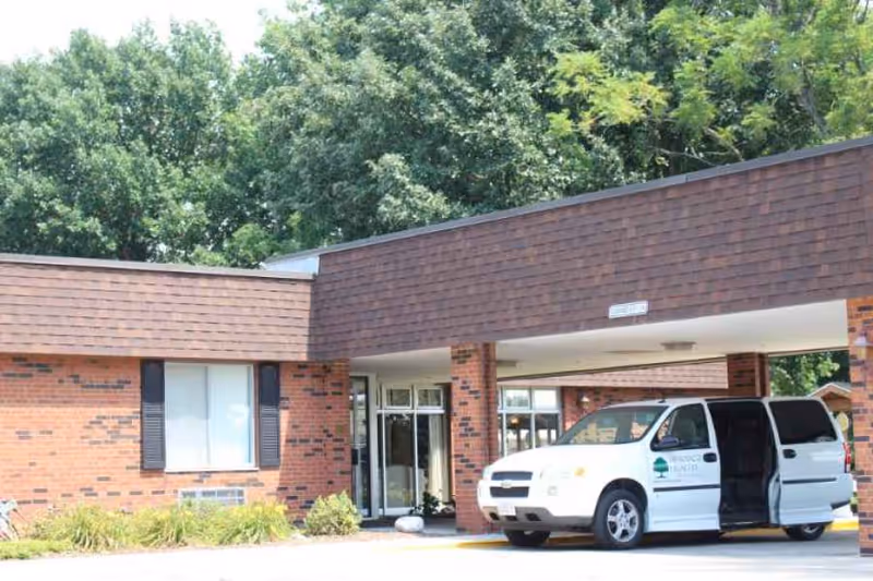 Exterior view of a brick building with a covered entrance. A white van with a sliding door open is parked in front of the entrance. There are trees and greenery in the background.