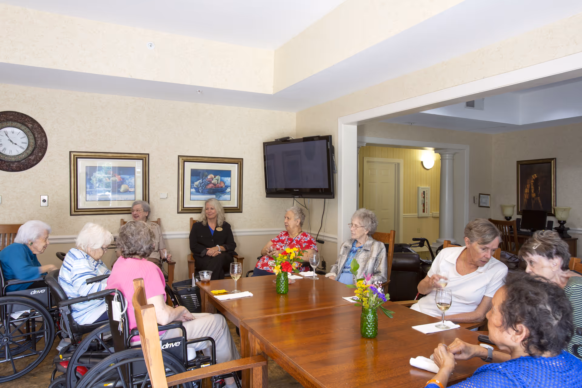 A group of elderly women sitting around a wooden table in a common room, some in wheelchairs, engaged in conversation. The room has beige walls with framed pictures, a wall clock, and a mounted flat-screen TV. There are small flower vases on the table and a doorway leading to another room in the background.