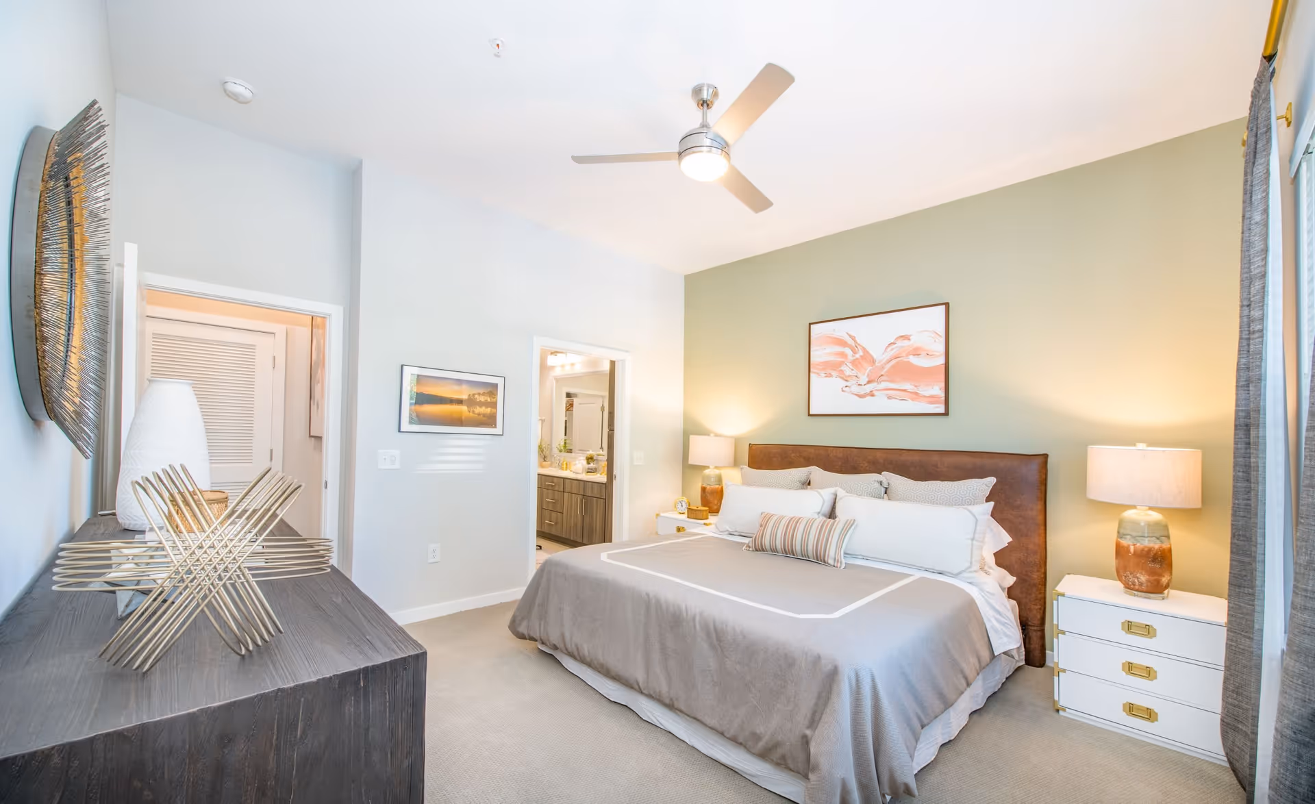 Bright, modern bedroom featuring a large bed with pillows, two bedside tables with lamps, a dresser with decorative objects, and a ceiling fan.