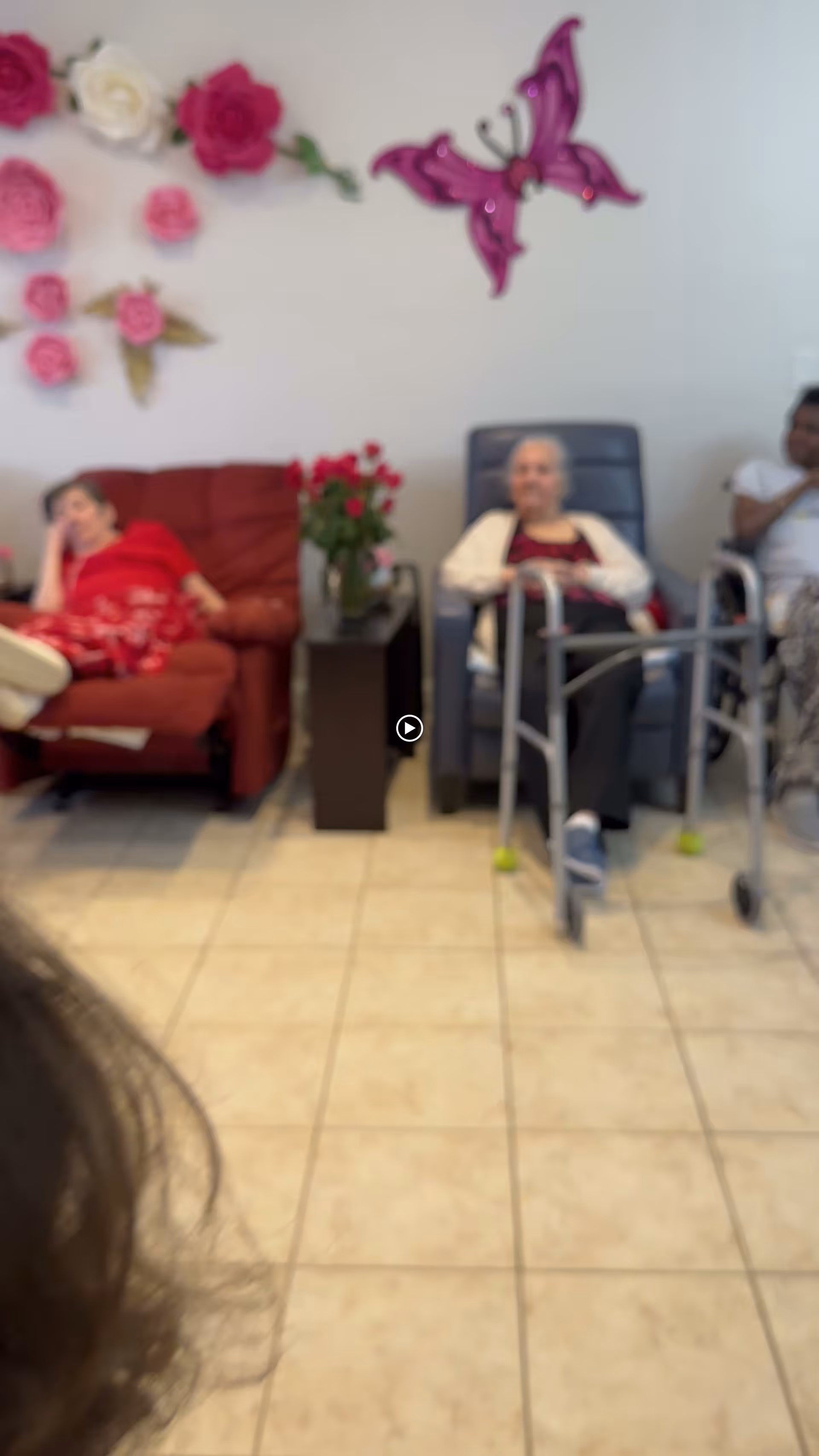 Three elderly individuals sitting in a living room area of an assisted living facility. One person is reclining on a red armchair, another is seated in a blue chair with a walker in front, and the third person is sitting on the right side. The wall behind them is decorated with large pink and white flower decorations and a purple butterfly. There is a small table with a vase of red flowers between the chairs.
