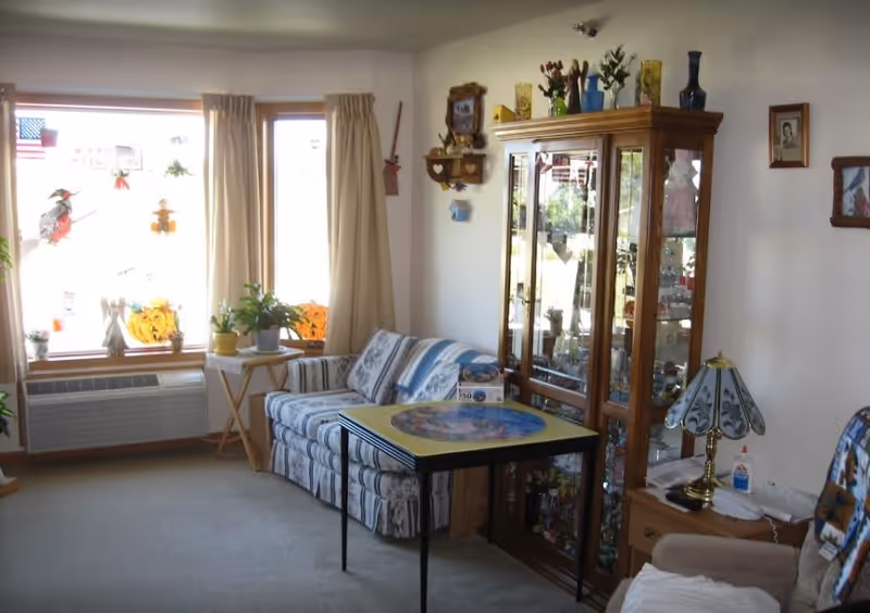 A cozy living room with a patterned couch, a small table with a puzzle on it, a wooden display cabinet filled with decorative items, a side table with a lamp and glue, and windows with beige curtains decorated with autumn and Halloween-themed stickers.