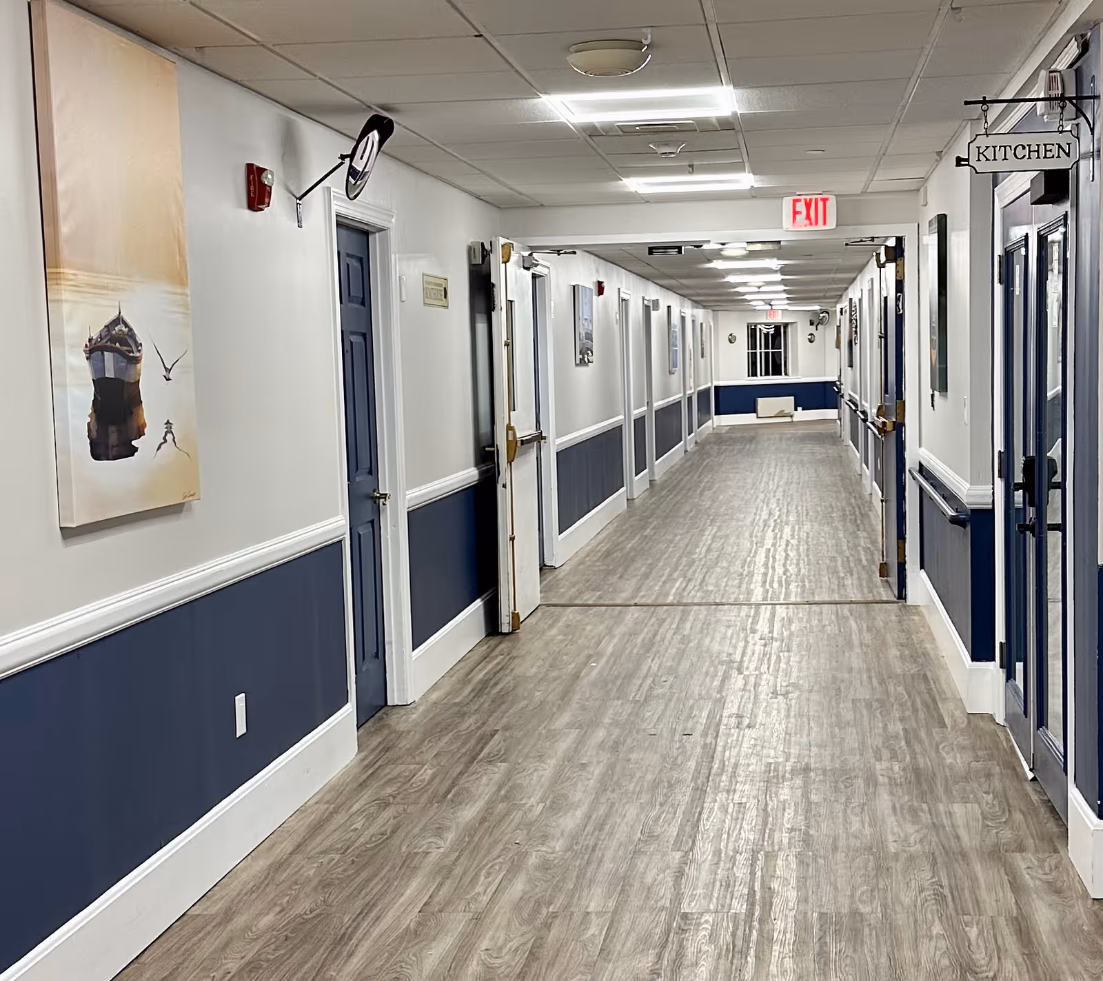 A long, well-lit hallway in an assisted living facility with wood-patterned flooring, white walls with blue wainscoting, and multiple closed doors on both sides. There is a sign labeled 'KITCHEN' hanging on the right side near a door, and an exit sign is visible at the far end of the corridor. A painting and a fire alarm are mounted on the left wall.