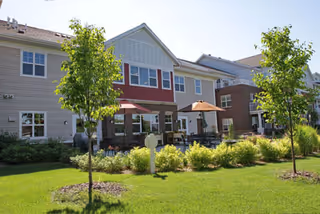 Exterior view of a senior living facility building with beige and red siding, multiple windows, and a patio area with tables and umbrellas. The foreground features a well-maintained lawn, small trees, and shrubs under a clear blue sky.