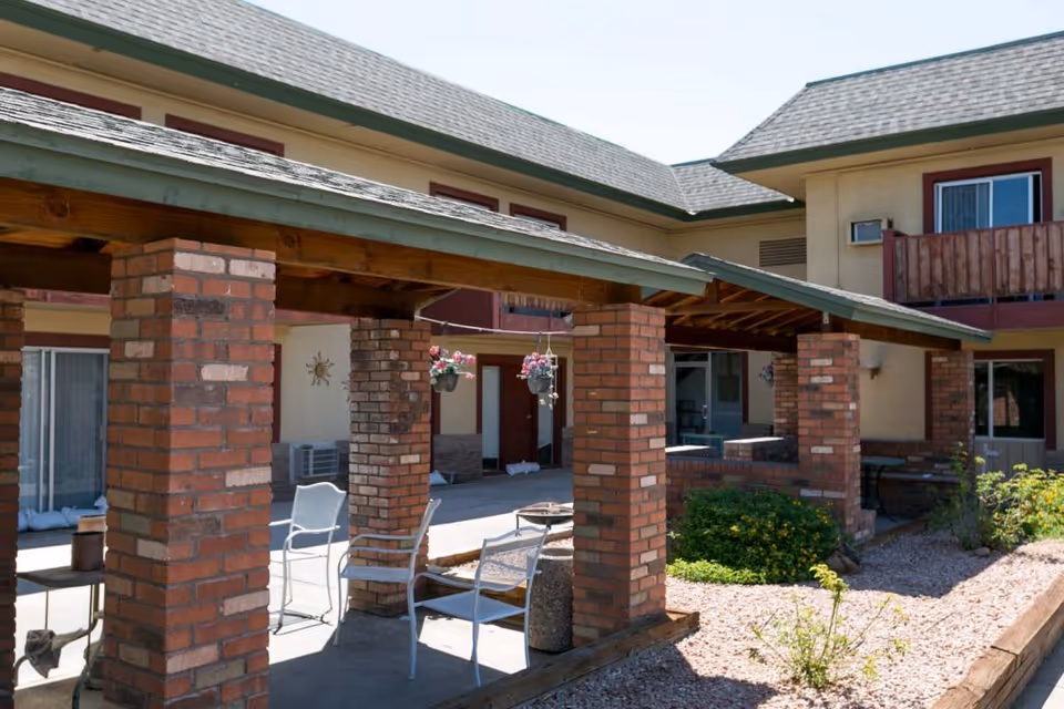 Outdoor patio area at Rose Court Senior Living with brick pillars supporting a wooden roof structure, white chairs, hanging flower pots, and surrounding garden beds with shrubs and gravel.