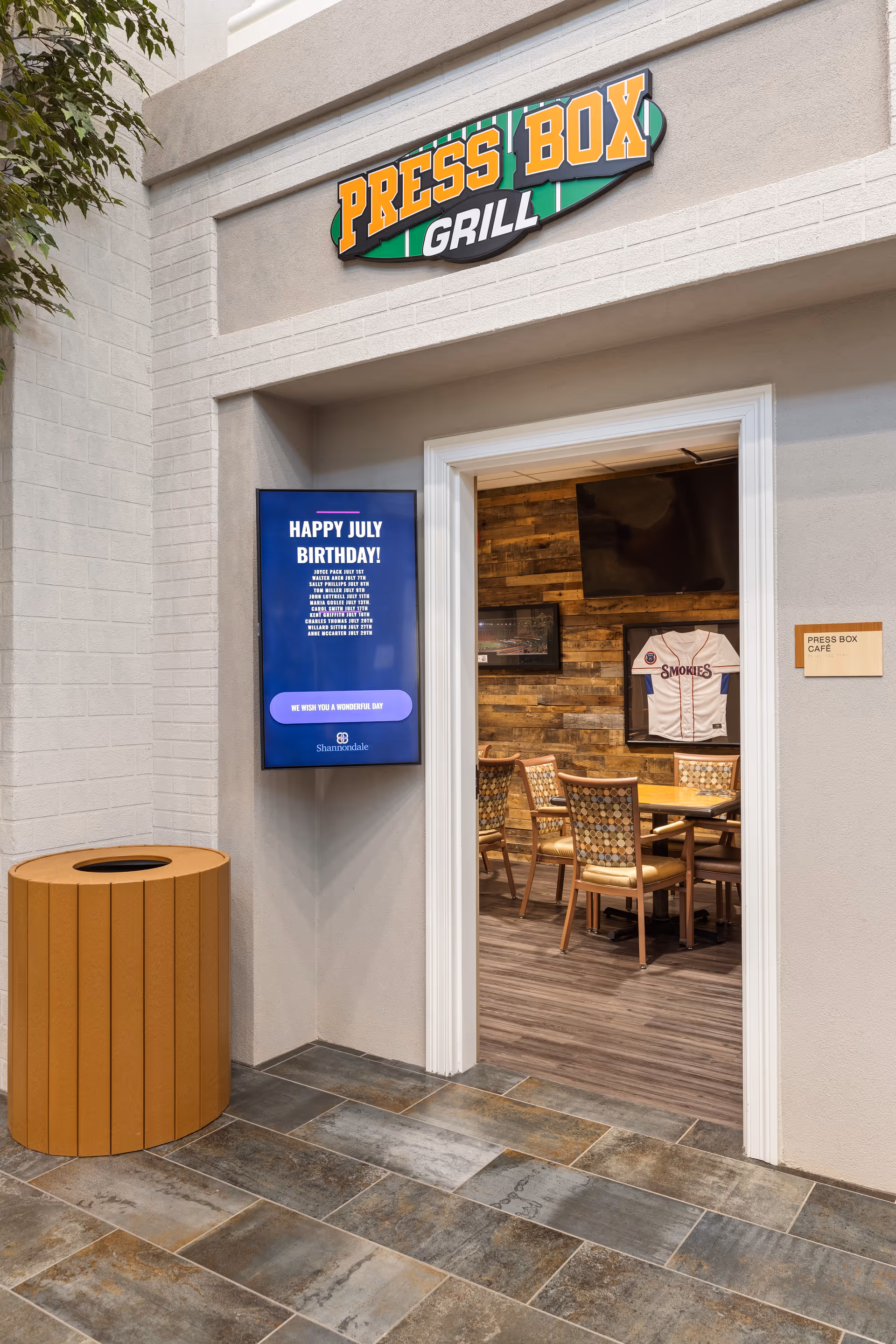 Entrance to the Press Box Grill at Shannondale of Knoxville, featuring a sign above the doorway, a digital display wishing happy July birthdays, a wooden trash can, and a view inside showing a table with chairs and a framed Smokies baseball jersey on a wooden accent wall.