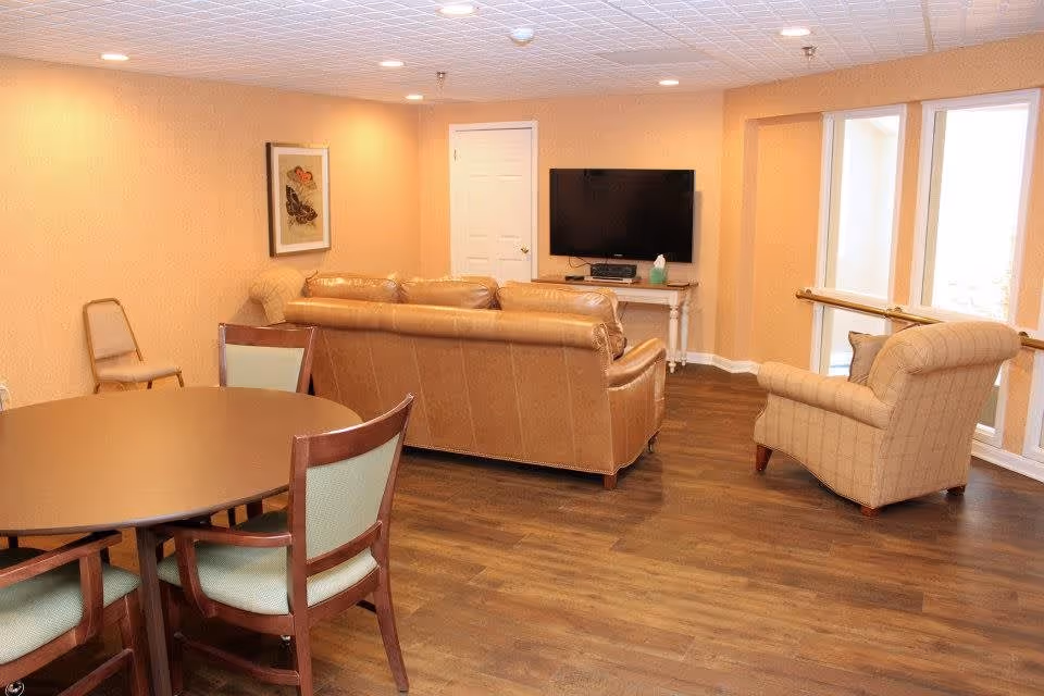 A cozy living room area with a round wooden table and four chairs in the foreground, a tan leather sofa facing a wall-mounted flat-screen TV, and a beige armchair near large windows letting in natural light. The room has wooden flooring, warm beige walls, recessed ceiling lights, and a framed picture on the wall.
