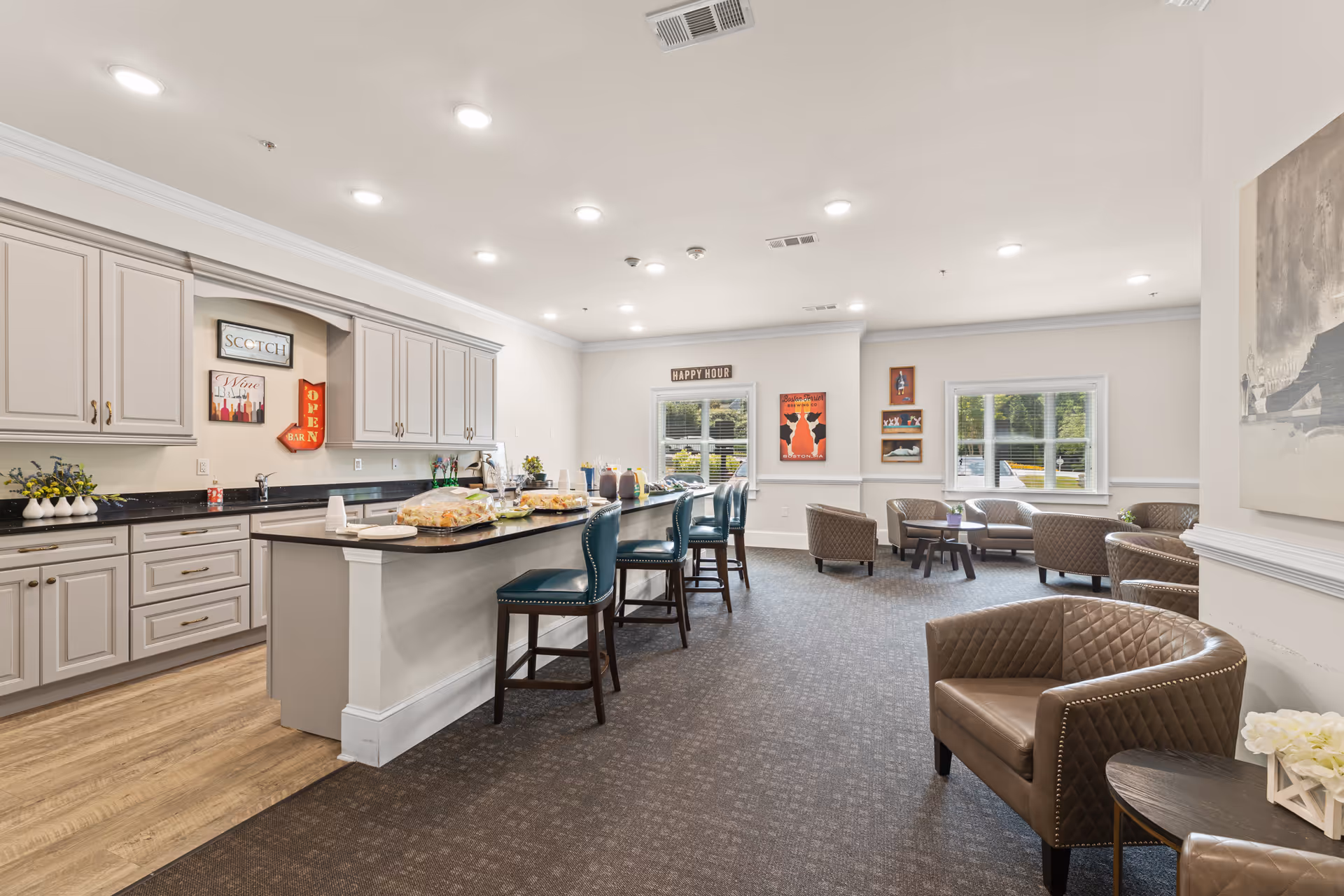 A spacious and well-lit common area featuring a kitchen with light gray cabinets, a black countertop island with teal bar stools, and a seating area with brown leather chairs and small tables. The walls are decorated with framed artwork and signs, including one that says 'HAPPY HOUR'. Large windows allow natural light to fill the room.
