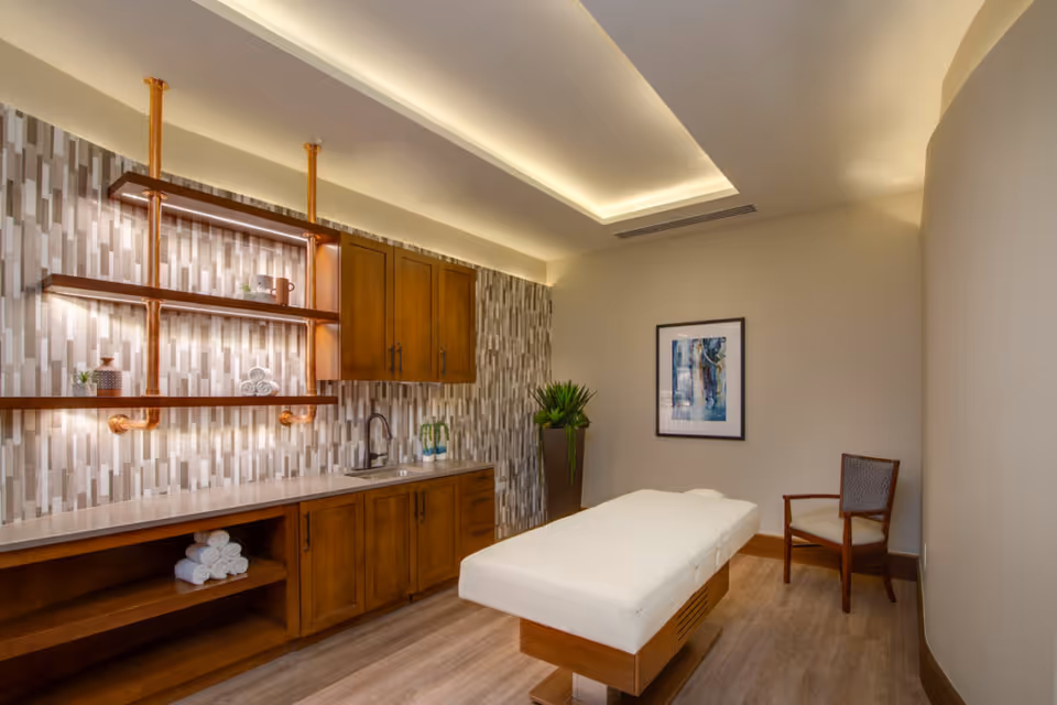 A serene massage or therapy room with a padded treatment table in the center, wooden cabinetry with a sink and shelves holding rolled towels and decorative items, a wooden chair, a potted plant, and a framed abstract artwork on the wall. The room features warm lighting and a modern ceiling design.