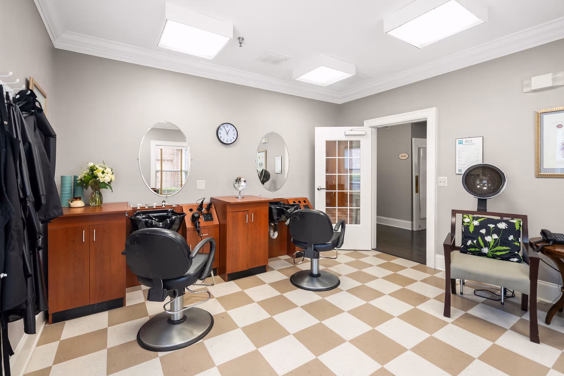 Interior of a hair salon area with two black salon chairs in front of wooden cabinets with sinks and oval mirrors. There are black salon capes hanging on hooks to the left, a clock on the wall, and a chair with a floral cushion next to a hair dryer on the right. The floor has a beige and white checkered pattern.