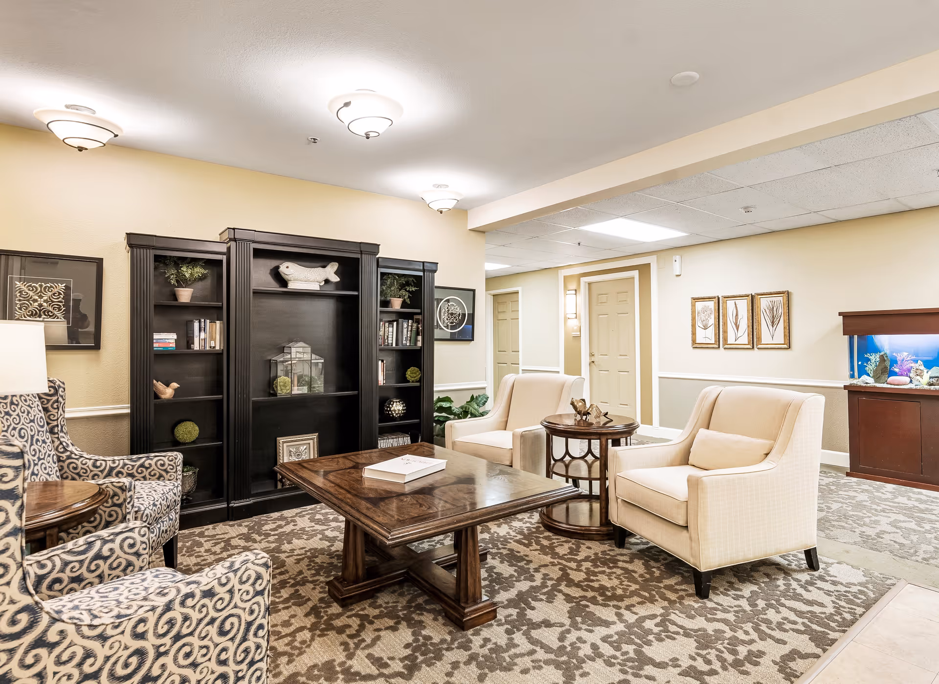 A cozy living room area in a senior living facility featuring two beige armchairs, two patterned armchairs, a large wooden coffee table, and a dark wooden bookshelf with decorative items and books. The room has soft yellow walls, framed artwork, ceiling lights, and a fish tank on a wooden stand in the background.