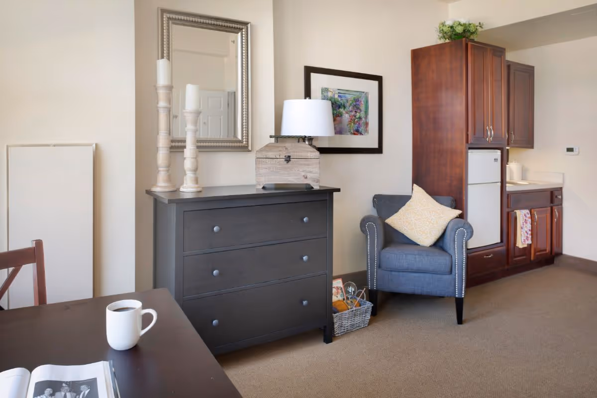 A cozy interior room featuring a dark wooden dresser with two tall white candles and a lamp on top, a framed mirror and colorful artwork on the wall, a blue armchair with a yellow patterned pillow, a small kitchenette with wooden cabinets and a mini fridge, and a table with a white coffee mug and an open book.