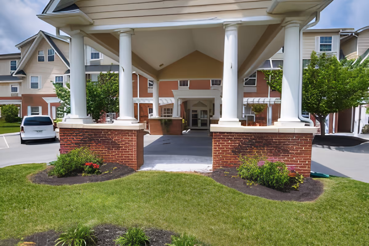 Covered front entrance/portico with white columns and brick bases at a senior living building, with landscaping and a parked vehicle nearby.