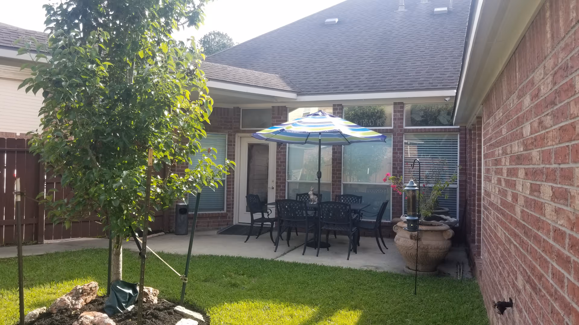 Outdoor patio area with a table and six chairs under a blue and green striped umbrella. The patio is surrounded by brick walls and windows, with a small tree and green grass in the foreground. There is a large decorative planter with plants and a bird feeder nearby.