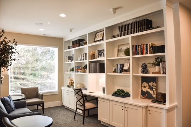 A cozy interior room with built-in white bookshelves filled with books, decorative items, and framed pictures. There is a wooden chair at a built-in desk area, a gray sofa with blue pillows, a small round table, and a large window with blinds letting in natural light.
