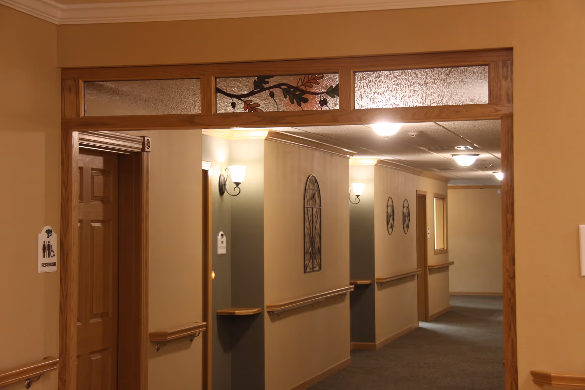 Interior hallway of a senior living facility with beige walls, wooden trim, handrails, wall sconces, decorative wall art, and a door marked as a restroom.