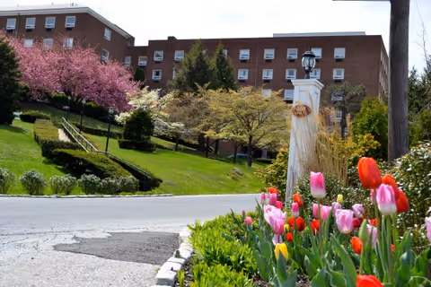 Brick senior living building set on a grassy landscaped hill with blooming trees and pink and red tulips in the foreground.