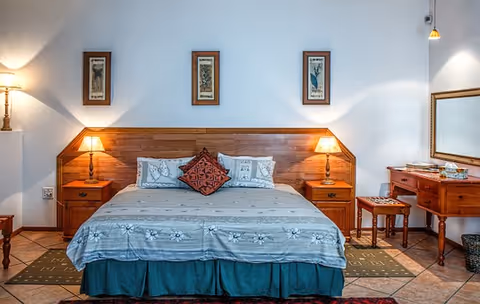 Well-lit bedroom with a large bed flanked by wooden nightstands and lamps, framed artwork above, and a dressing table with mirror to the right.