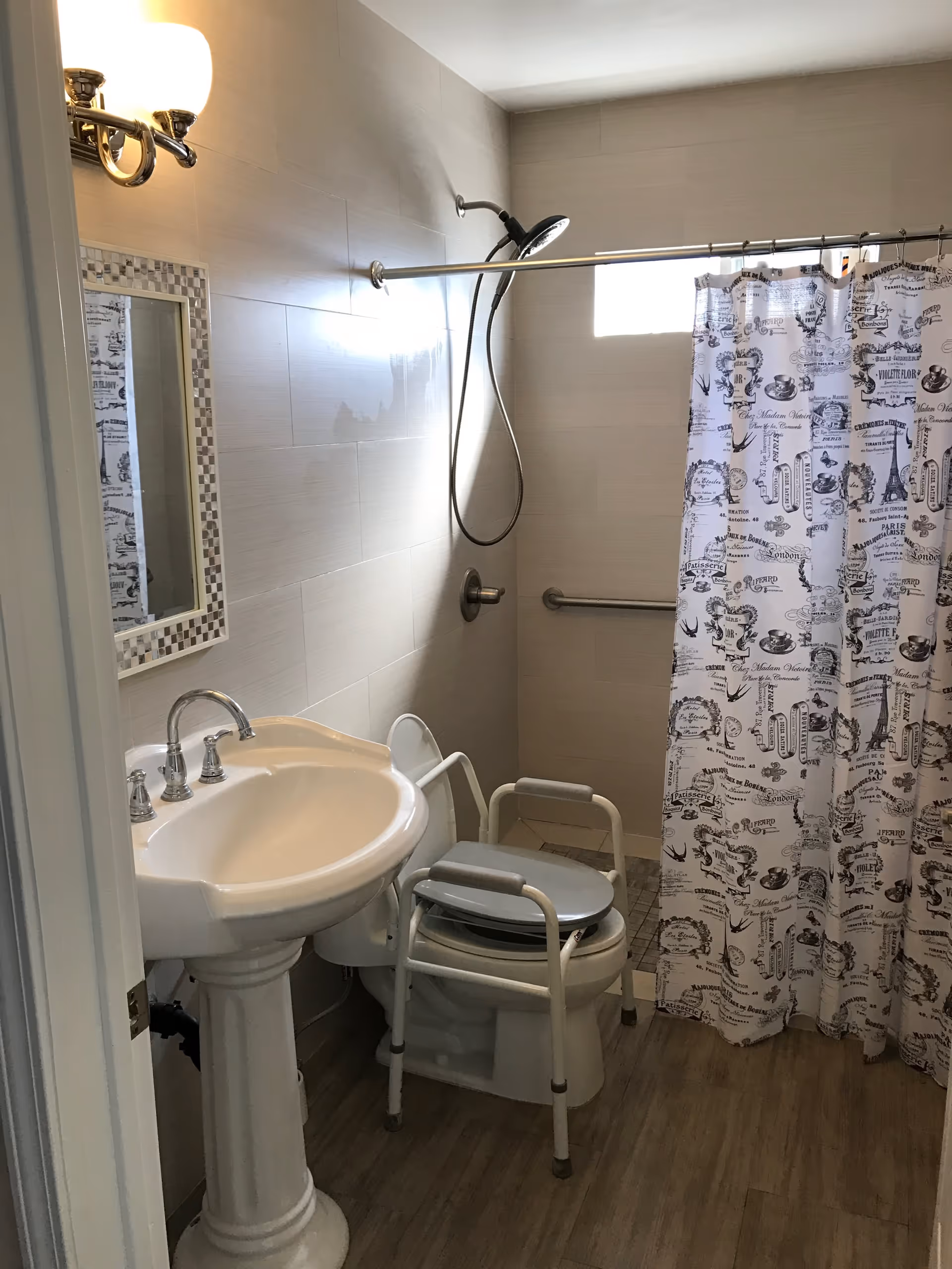 A small bathroom with a pedestal sink, a toilet with a raised seat and armrests for accessibility, a shower area with a handheld showerhead, a grab bar on the wall, and a shower curtain with black and white vintage-style text and images.