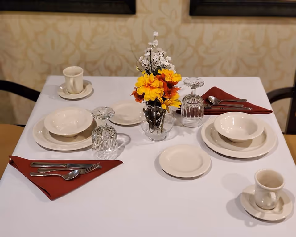 A dining table set for two with white plates, bowls, cups, and saucers, two upside-down clear glasses, silverware placed on red napkins, and a small vase with yellow and orange flowers in the center. The table is covered with a white tablecloth and has a patterned beige wallpaper background.