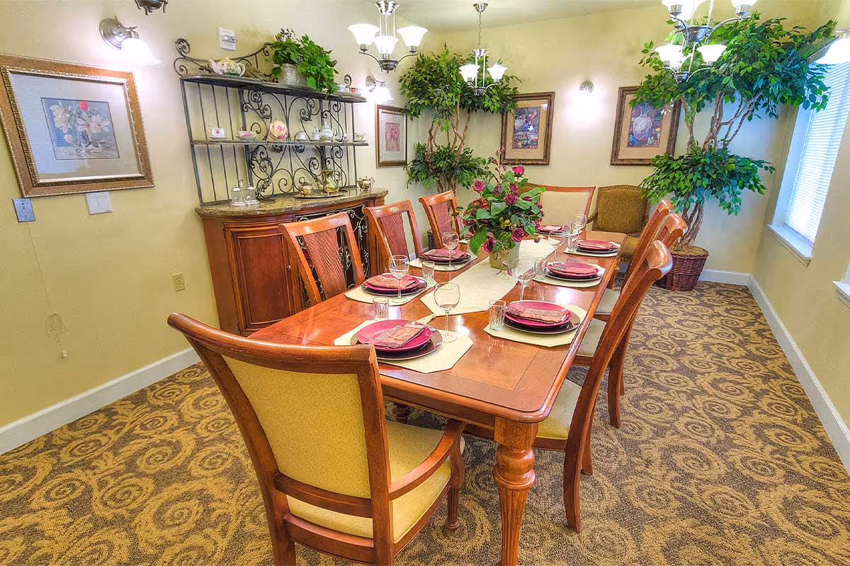 A well-lit dining room with a long wooden table set for eight people, featuring plates, glasses, and napkins. The room has patterned carpet, framed artwork on the walls, a decorative cabinet with shelves holding teapots and cups, and several large potted plants near the windows.