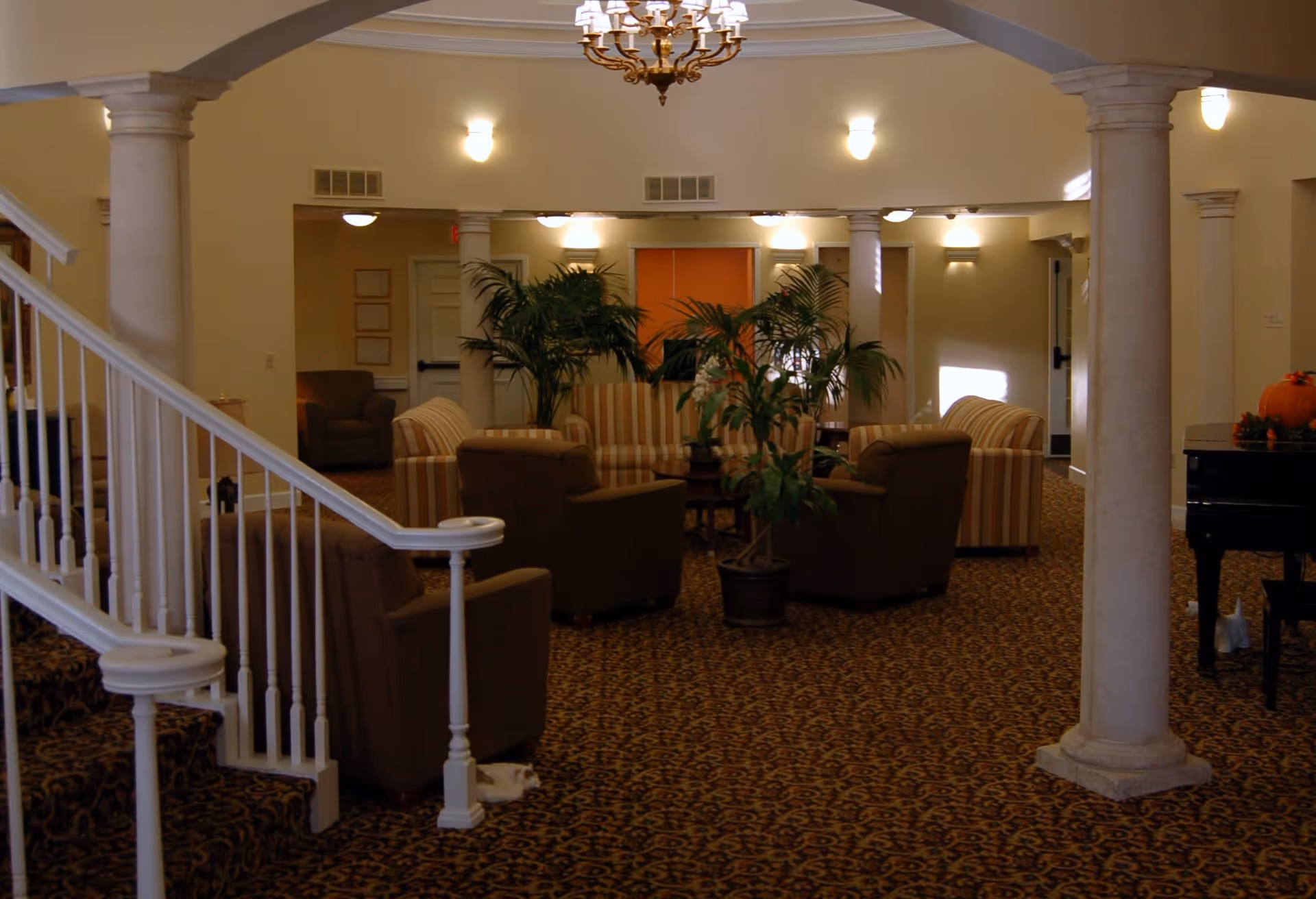 Interior view of a senior living facility lounge area with patterned carpet, several armchairs and striped sofas arranged around a central coffee table with potted plants. The room features white columns, a staircase with white railing on the left, a chandelier hanging from the ceiling, and warm lighting on the walls. A piano with a pumpkin decoration is visible on the right side.
