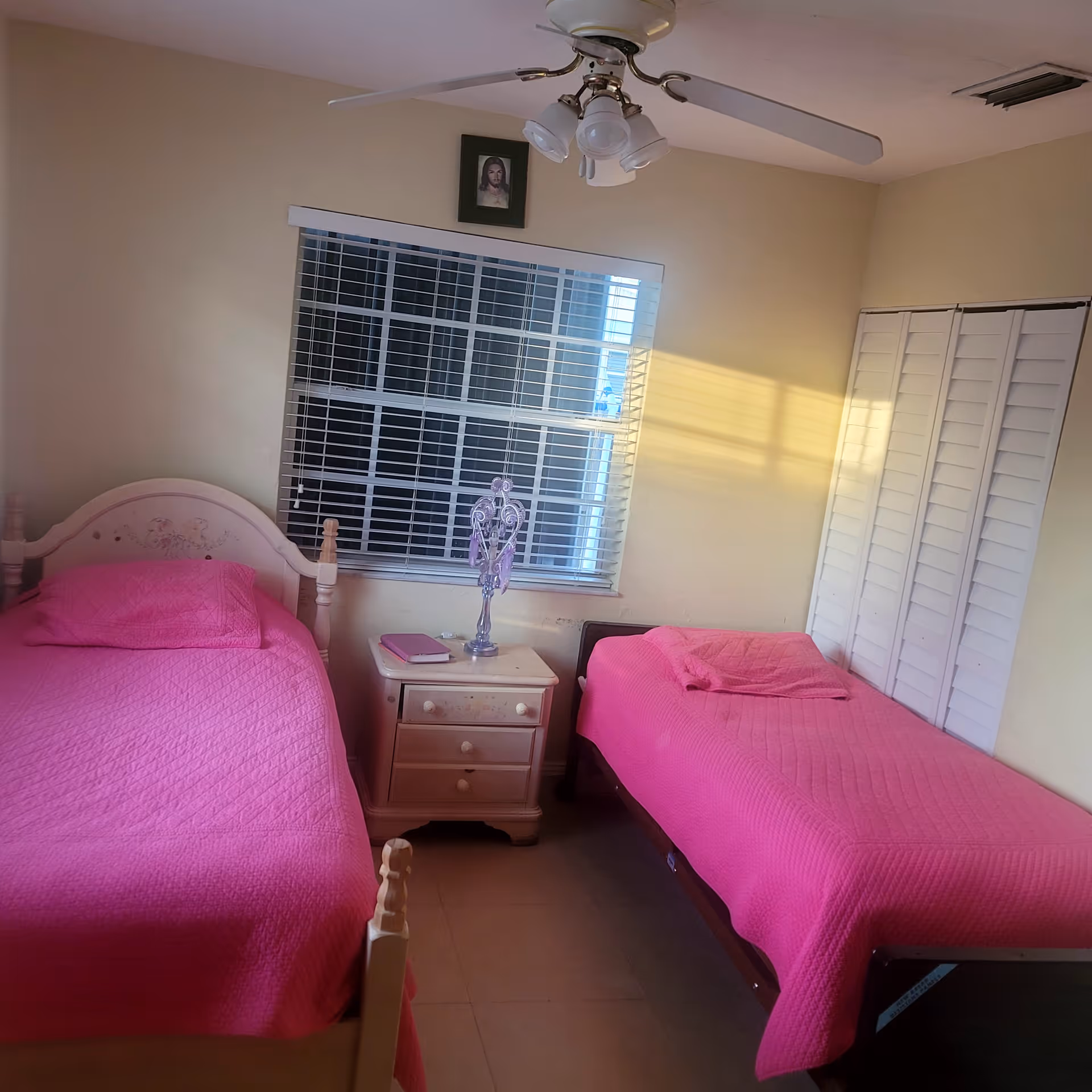 Small bedroom with two twin beds covered in bright pink quilts, a nightstand between them, and a window with blinds.