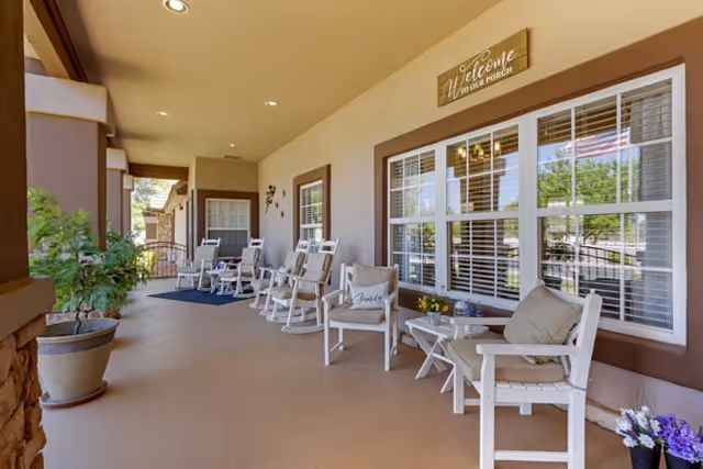A spacious covered porch area with several white rocking chairs and small tables arranged along the wall. There are potted plants and flowers placed around, and a wooden sign above the window reads 'Welcome to our porch.' The porch overlooks a green outdoor area visible through the windows.