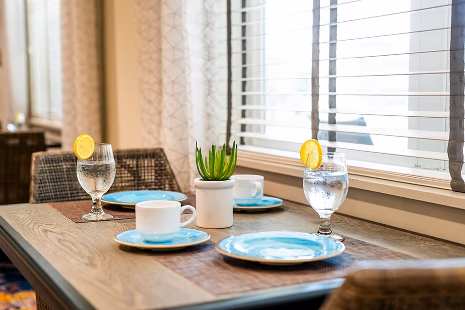 A dining table set for two with blue plates, white cups on saucers, and glasses of water garnished with lemon slices. A small potted plant is placed in the center of the table. The table is near a window with white blinds and patterned curtains.