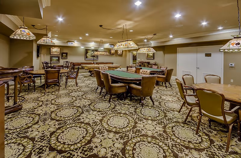 A spacious game room with multiple tables and chairs, including a poker table and a pool table. The room is decorated with patterned carpet, hanging stained glass lamps, and framed artwork on the walls. The lighting is warm and the ceiling has recessed lights.
