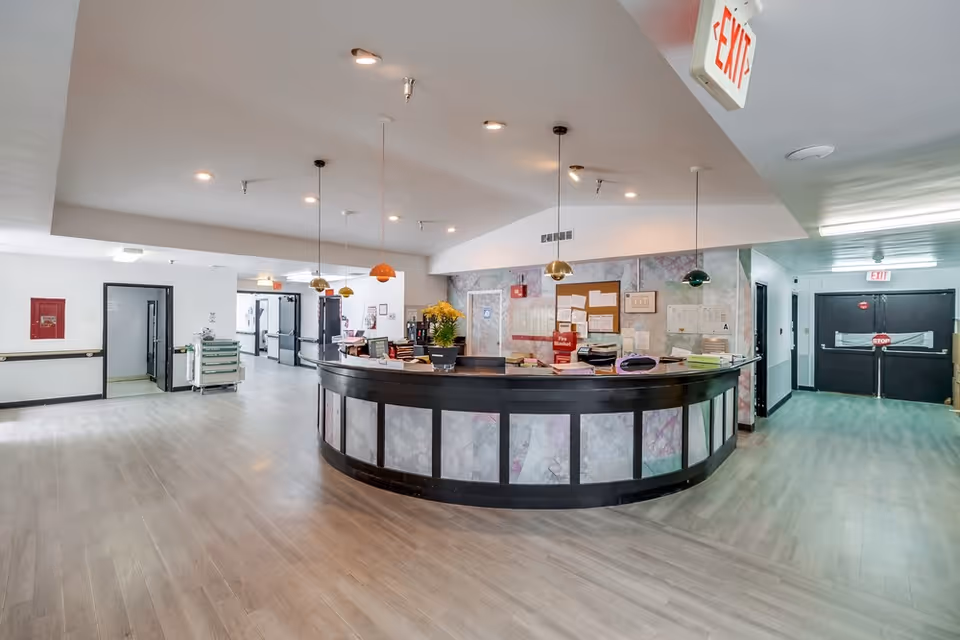 Interior view of a healthcare facility reception area with a curved front desk, overhead pendant lights in various colors, bulletin boards with notices on the wall behind the desk, and exit signs visible on the ceiling. The floor is light-colored wood, and there are doors and hallways leading to other parts of the facility.